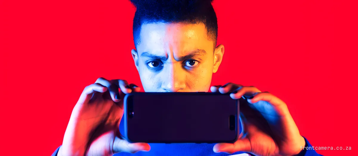A young South African creator filming themselves on a phone propped against a bright window, an open notebook with a numbered list on the sill, determined expression