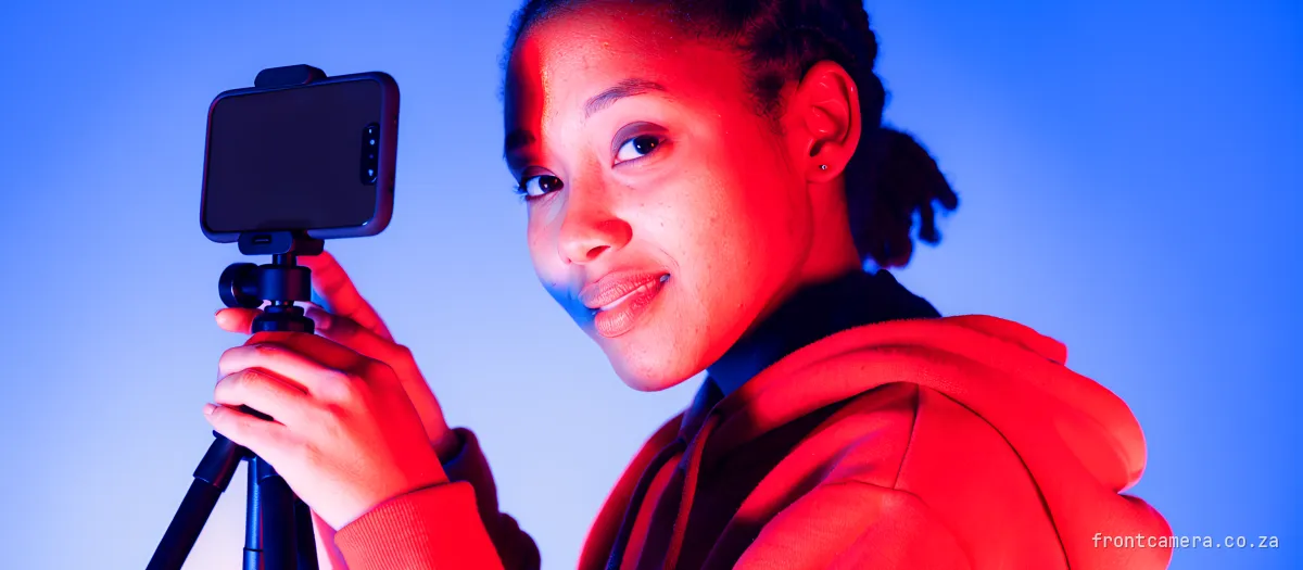 A young South African woman creator on a seaside promenade holding a phone on a small tripod, looking over her shoulder with a cautious half-smile